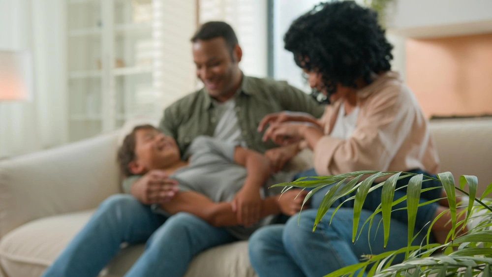 Bonding family african american parents with little adopted kid boy son tickling on home sofa