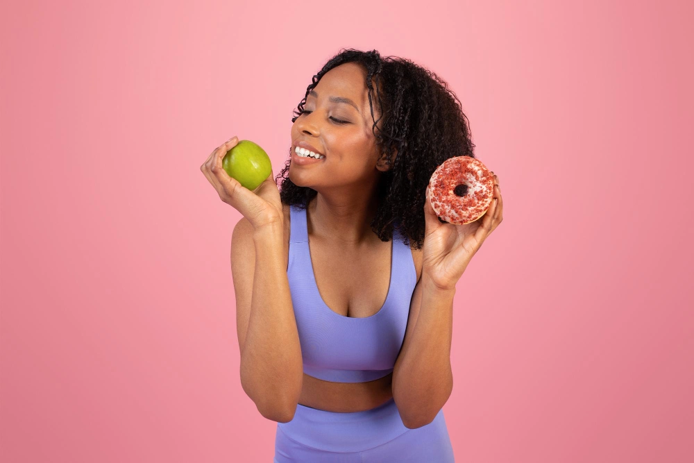 Cheerful pensive millennial African American lady in sportswear chooses donut or green apple