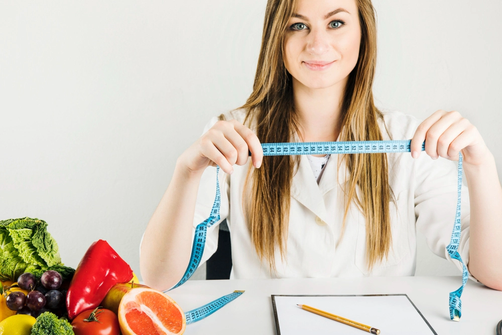 Smiling female dietician holding measuring tape with healthy food on desk