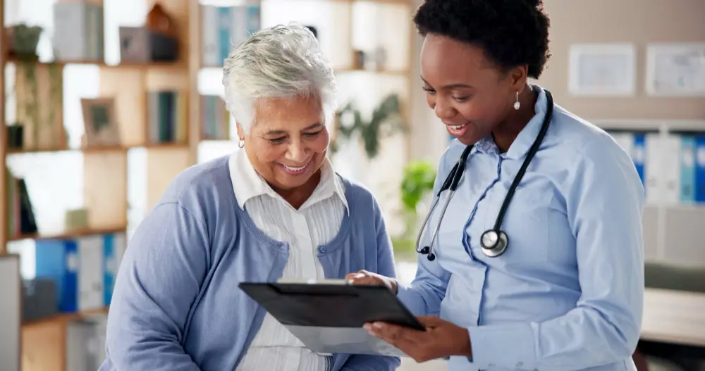 A woman in her 30s–50s talking with a clinician in scrubs or a white coat, both seated, with a clipboard or tablet visible.